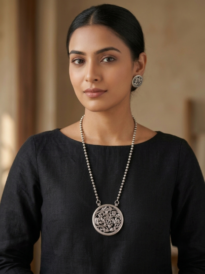 A woman wearing a silver necklace with a circular filigree pendant and matching earrings, dressed in a black top against a warm beige background.