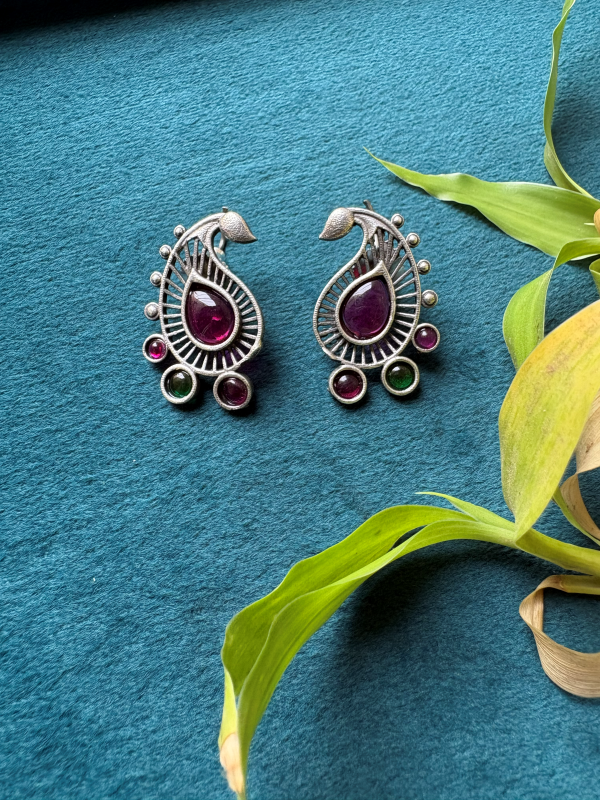 A pair of silver-toned paisley earrings with deep purple central stones and small green and purple accent stones, displayed on a textured teal surface with green leaves in the corner.