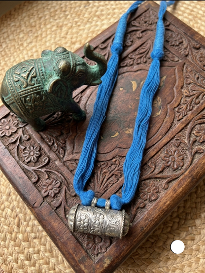 A blue thread necklace with a carved silver cylindrical pendant, displayed on a carved wooden box next to a small green elephant figurine.
